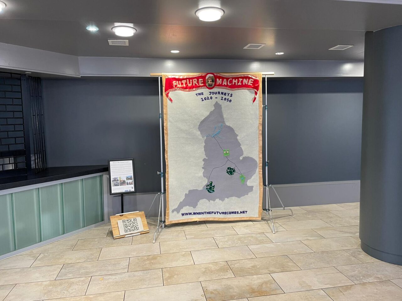 Textile banner with Future Machine the journeys written on it and a large map of England with symbols representing each of the of places where Future Machine appears on its journey, next to the banner is a poster on a stand and on the floor is a a clay QR code