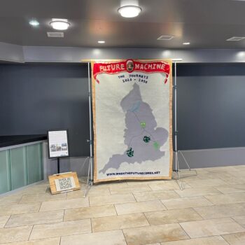 Textile banner with Future Machine the journeys written on it and a large map of England with symbols representing each of the of places where Future Machine appears on its journey, next to the banner is a poster on a stand and on the floor is a a clay QR code