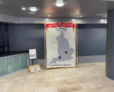 Textile banner with Future Machine the journeys written on it and a large map of England with symbols representing each of the of places where Future Machine appears on its journey, next to the banner is a poster on a stand and on the floor is a a clay QR code