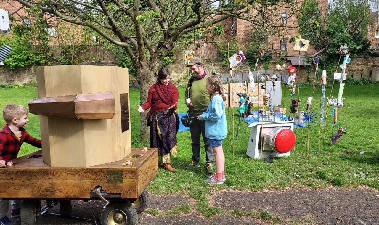 A child at the back of Future Machine, the Future Machine's companion talking to a man and a girl, with a blossom tree and objects on sticks made of recycled rubbish behind it and a large red button sitting on the grass.