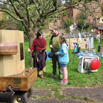 A child at the back of Future Machine, the Future Machine's companion talking to a man and a girl, with a blossom tree and objects on sticks made of recycled rubbish behind it and a large red button sitting on the grass.