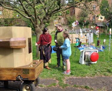 A child at the back of Future Machine, the Future Machine's companion talking to a man and a girl, with a blossom tree and objects on sticks made of recycled rubbish behind it and a large red button sitting on the grass.