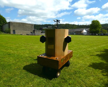 Future Machine in the rugby field with blue sky and clouds above, The Lakes School, and a wooded hill behind