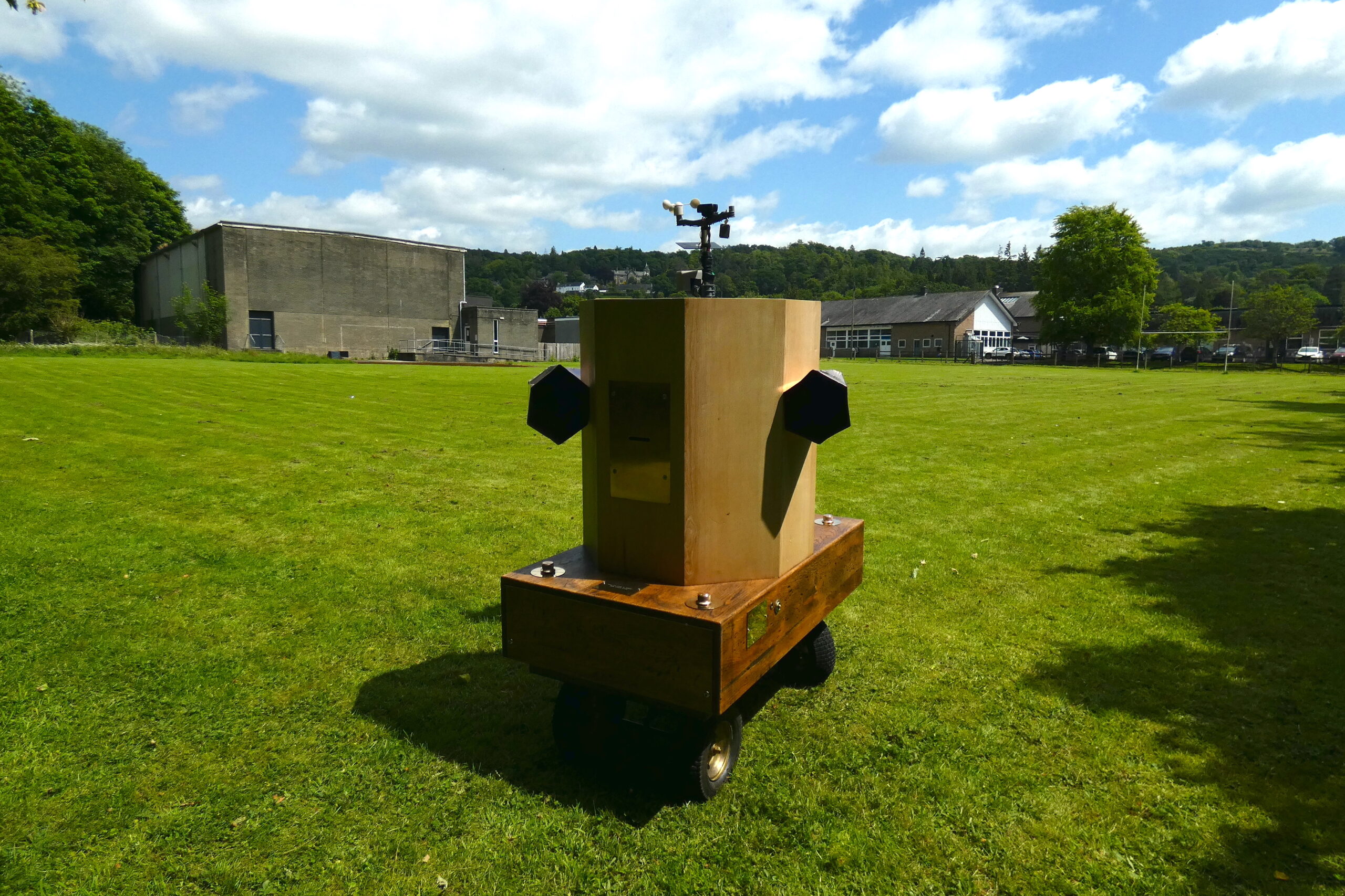 Future Machine in the rugby field with blue sky and clouds above, The Lakes School, and a wooded hill behind