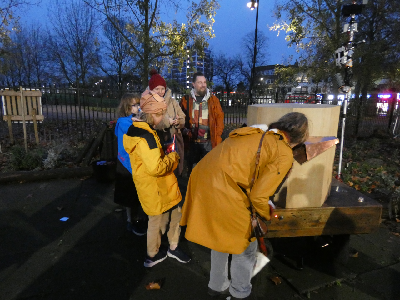 a family speaking to Future Machine with the park and buidings in the background, at dusk