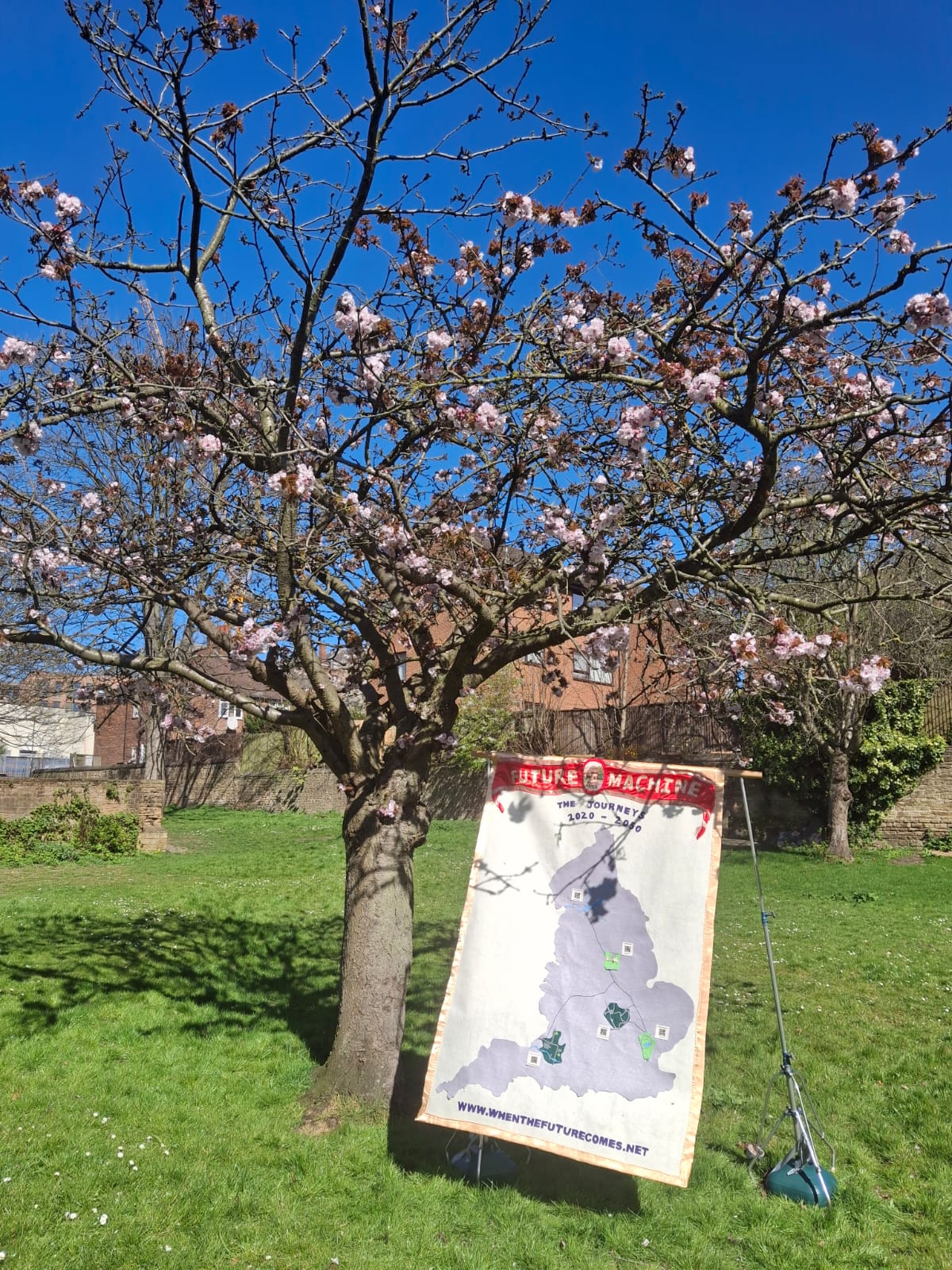A blossom tree with the top of a large textile banner underneath with the words Future Machine The Journeys 2020-2050 embroidered on it.