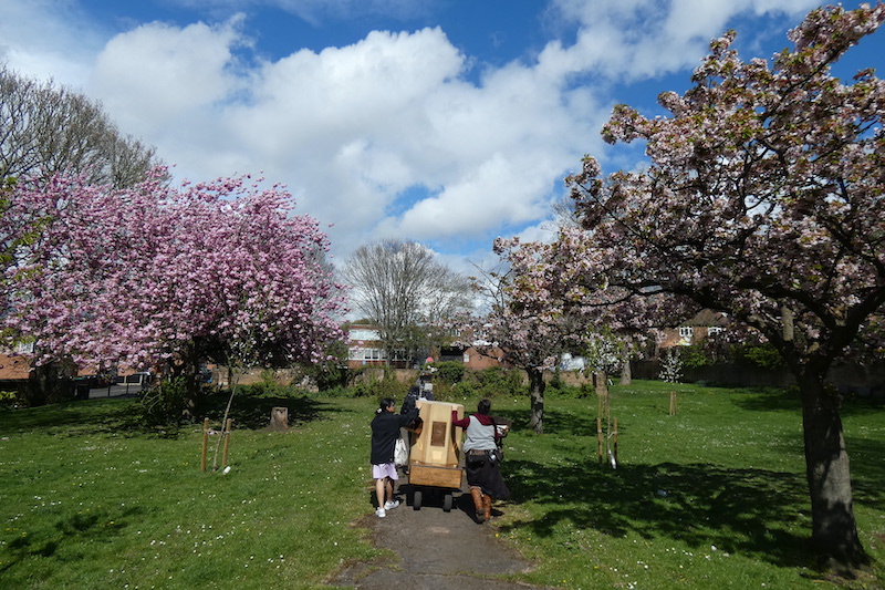 Future Machine's companion and Sanjana pushing Future Machine along a path with blossom trees on either side