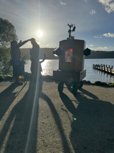 Future Machine with Lake Windermere behind, a low sun glinting on the water and 3 people with around them