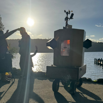 Future Machine with Lake Windermere behind, a low sun glinting on the water and 3 people with around them
