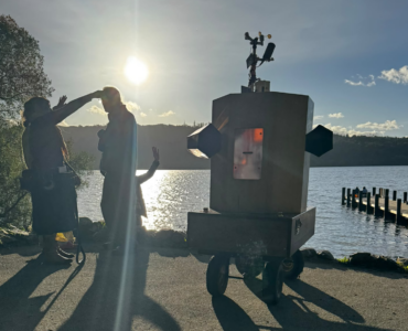 Future Machine with Lake Windermere behind, a low sun glinting on the water and 3 people with around them