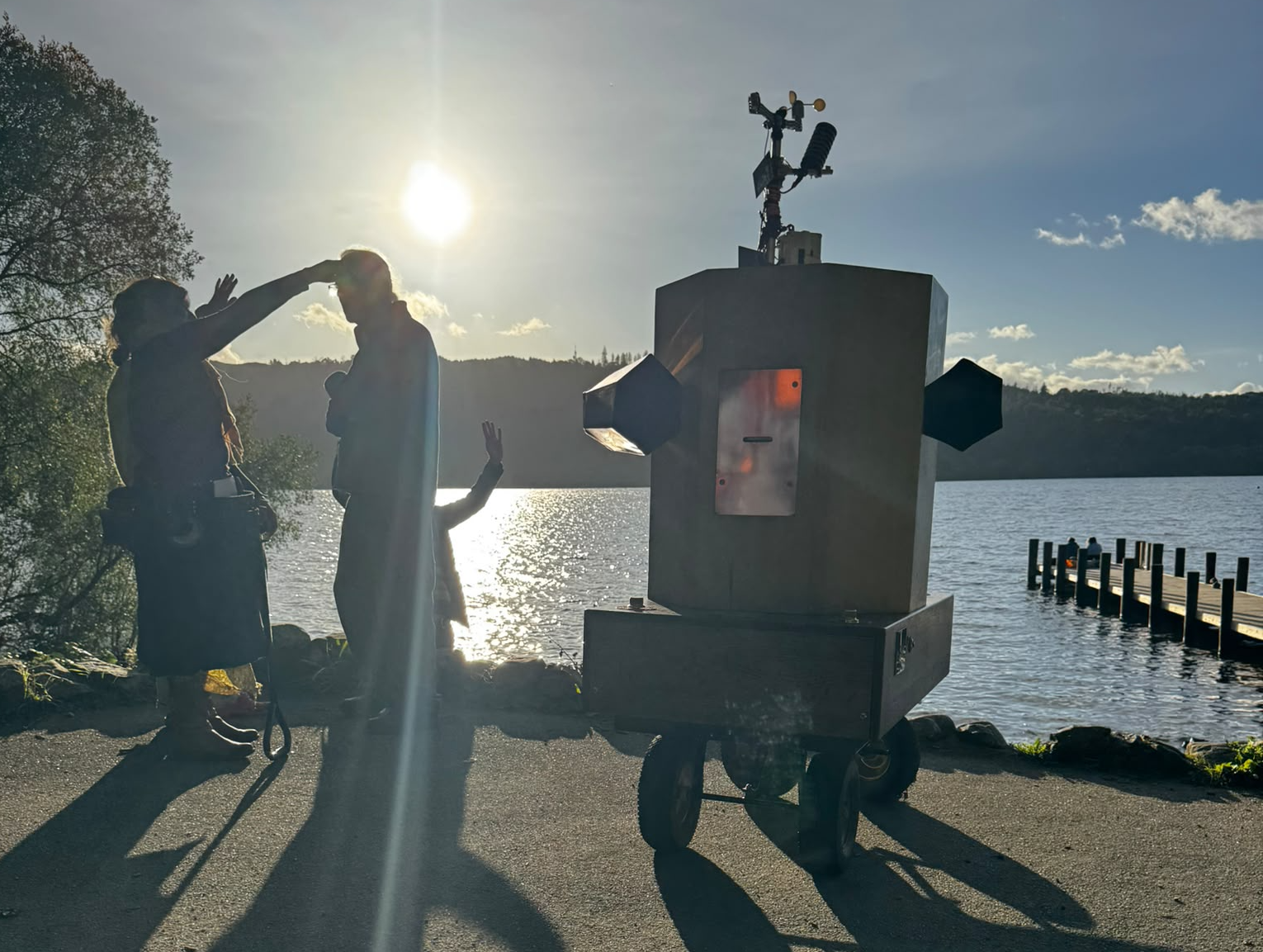 Future Machine with Lake Windermere behind, a low sun glinting on the water and 3 people with around them